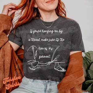May include: A dark gray t-shirt with a white graphic design featuring a line drawing of a hand reaching for the hem of a garment. The text "If you're hanging on by a thread, make sure it's the hem of His garment." is printed on the shirt. The scripture reference "Matthew 14:20-22" is printed below the graphic.