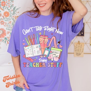 May include: Purple t-shirt with the text "Can't Talk Right Now Doing Teacher Stuff" in rainbow letters. The shirt features a graphic of school supplies including a pencil, ruler, scissors, and a notebook.