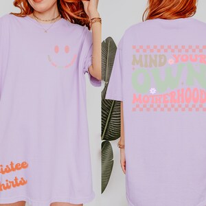 May include: A lavender t-shirt with a smiley face on the front and the text "Mind Your Own Motherhood" on the back. The text is in a green and pink checkered pattern.