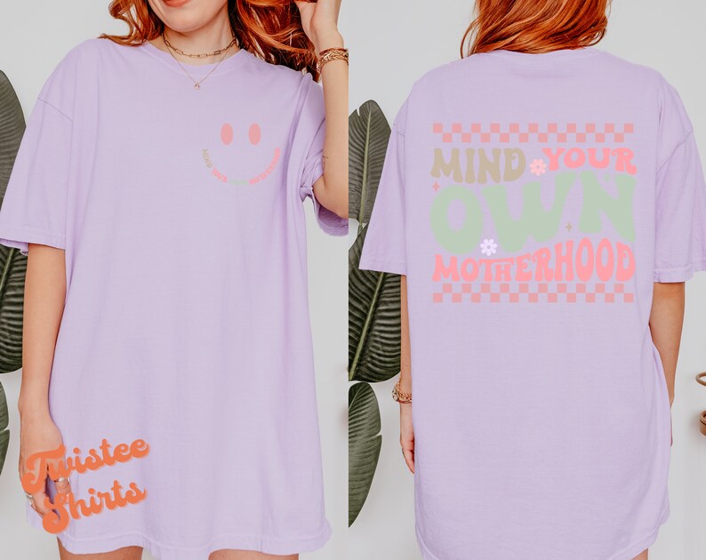 May include: A lavender t-shirt with a smiley face on the front and the text "Mind Your Own Motherhood" on the back. The text is in a green and pink checkered pattern.