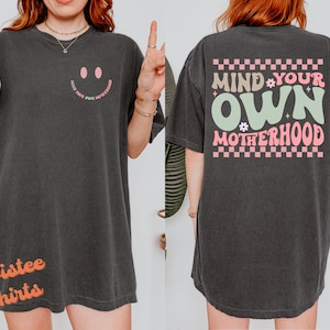 May include: Black oversized t-shirt with a pink smiley face on the front and the text "Mind Your Own Motherhood" in pink and green on the back. The shirt has a checkered pattern around the text.