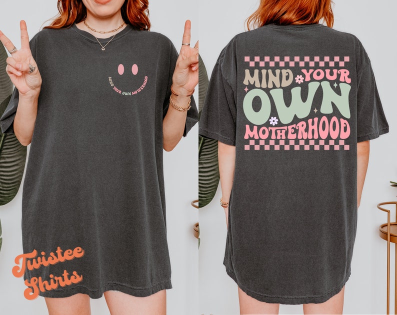 May include: Black oversized t-shirt with a pink smiley face on the front and the text "Mind Your Own Motherhood" in pink and green on the back. The shirt has a checkered pattern around the text.