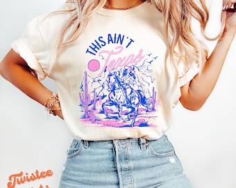 May include: A white t-shirt with a blue and pink graphic design featuring a woman riding a horse in a desert landscape. The text "This Ain't Texas" is printed on the shirt.