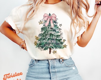 May include: A white t-shirt with a watercolor illustration of a Christmas tree with a pink bow and the text "Oh Come Let Us Adore Him".