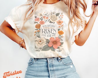 Pode incluir: Uma camiseta branca com um design floral e o texto "Ele não está aqui, pois ressuscitou, como disse".