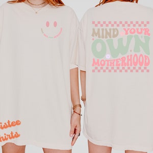 May include: A white t-shirt with a pink smiley face on the front and the text "Mind Your Own Motherhood" in pink and green on the back. The back design also features a checkered pattern in pink and white.
