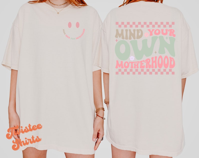 May include: A white t-shirt with a pink smiley face on the front and the text "Mind Your Own Motherhood" in pink and green on the back. The back design also features a checkered pattern in pink and white.