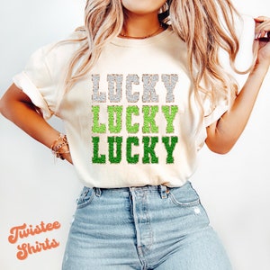 May include: A white t-shirt with the word "LUCKY" written three times in green and silver glitter. The shirt is being worn by a person with long blonde hair.