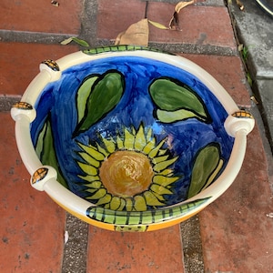 May include: A white ceramic bowl with a blue, green, and yellow sunflower design. The bowl has a scalloped rim and a handle on each side.