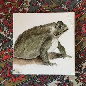 May include: A hand-painted ceramic tile featuring a detailed illustration of a green toad with a white belly. The toad is sitting on a light brown background. The tile is signed "Ju 1/92" in the bottom left corner.
