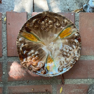May include: A brown and white ceramic plate with a raised design of grapes and leaves. The plate has a swirl pattern in the glaze, with shades of brown, yellow, and white.