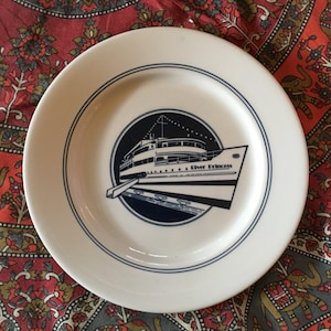 May include: A white ceramic plate with a blue border and a blue and white illustration of a ship with the text "River Princess" on the side.