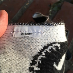 Op de afbeelding: Close-up van een wit-zwarte gebreide sjaal met een wit label waarop "Tim Burton's" staat.