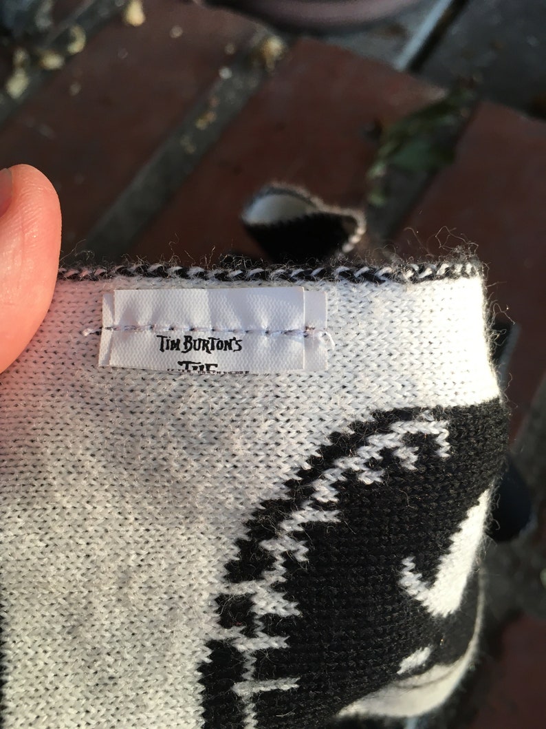 Op de afbeelding: Close-up van een wit-zwarte gebreide sjaal met een wit label waarop "Tim Burton's" staat.