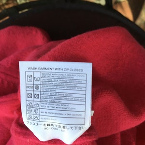 May include: A red care label with white text and symbols. The label includes instructions for washing, drying, ironing, and dry cleaning a garment. The text reads "Wash garment with zip closed" and "ファスナーを閉めて洗濯して下さい".