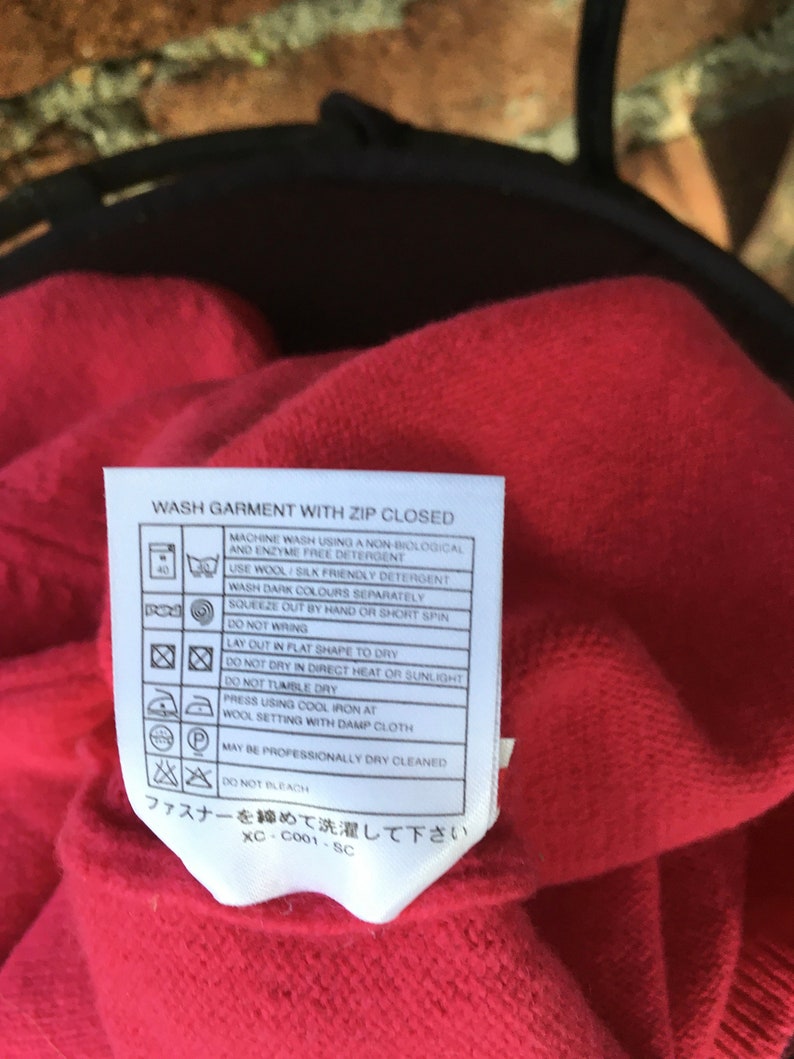 May include: A red care label with white text and symbols. The label includes instructions for washing, drying, ironing, and dry cleaning a garment. The text reads "Wash garment with zip closed" and "ファスナーを閉めて洗濯して下さい".