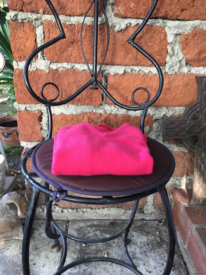 May include: A pink sweater folded and placed on a black metal chair with a brown cushion. The chair is in front of a brick wall.