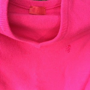 May include: A bright pink sweater with a small embroidered logo on the front. The sweater has a V-neck and is made of a soft, knit fabric.