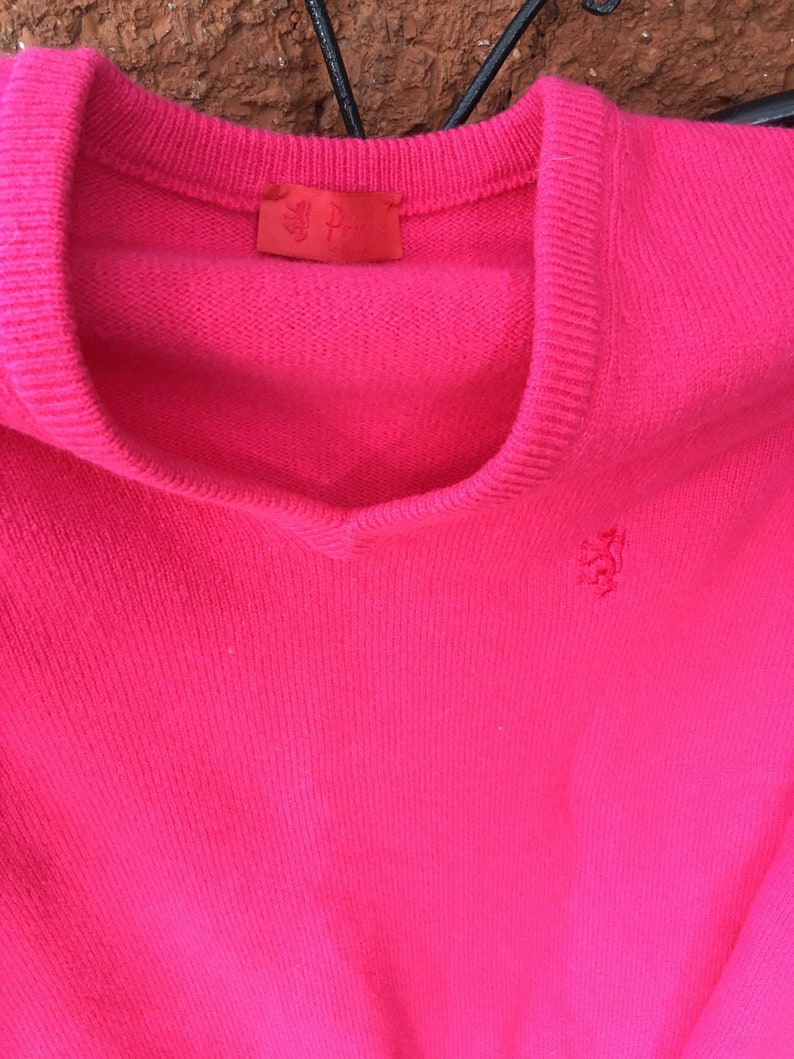 May include: A bright pink sweater with a small embroidered logo on the front. The sweater has a V-neck and is made of a soft, knit fabric.
