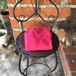 May include: A pink knitted sweater with a V-neck sits on a black metal chair with a brown cushion. The chair is in front of a brick wall.