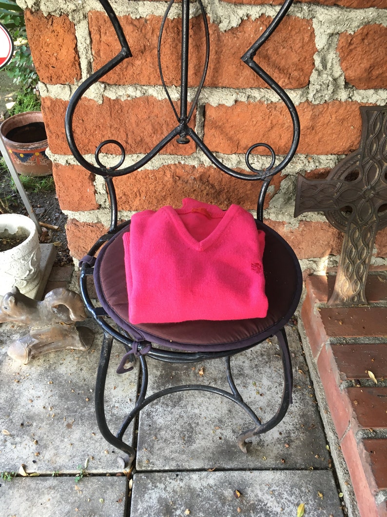 May include: A pink knitted sweater with a V-neck sits on a black metal chair with a brown cushion. The chair is in front of a brick wall.