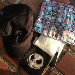 Op de afbeelding: Een zwart-witte gebreide sjaal met een zwart-witte afbeelding van Jack Skellington uit The Nightmare Before Christmas. Een zwarte pet met een rood en wit geborduurd ontwerp en een blauwe en paarse stoffen tas met een herhalend patroon van Jack Skellington en Sally uit The Nightmare Before Christmas.