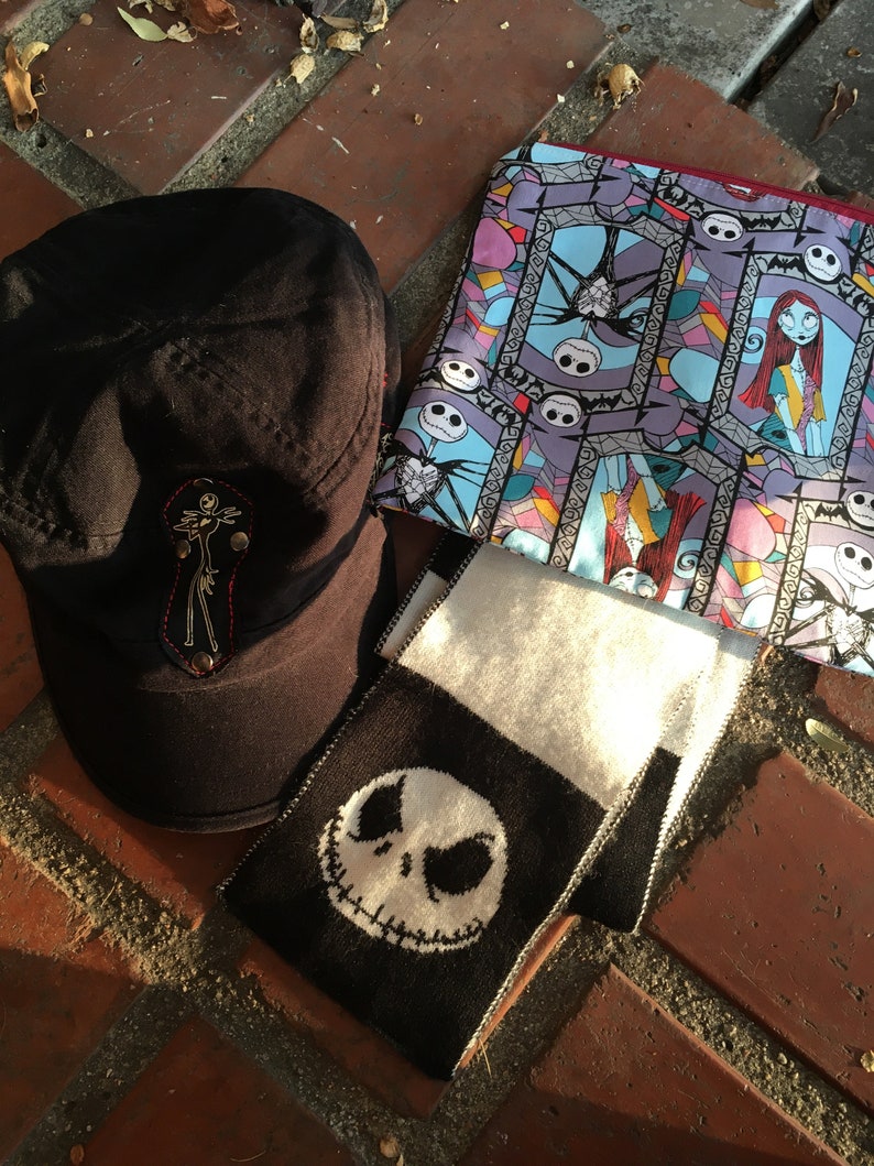 Op de afbeelding: Een zwart-witte gebreide sjaal met een zwart-witte afbeelding van Jack Skellington uit The Nightmare Before Christmas. Een zwarte pet met een rood en wit geborduurd ontwerp en een blauwe en paarse stoffen tas met een herhalend patroon van Jack Skellington en Sally uit The Nightmare Before Christmas.