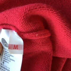 May include: A white tag with black text attached to a red sweater. The tag says "WOOLN", "LAMBSW", "LAINE D'AGNEAU", "LA VIRGEM", "LANA DE CORDERO", "LAMMWOLLE", "LAMMEULL", "毛", "(株)サン・フレール", "大阪市西区阿波座 1-4-16", "DE IN SCOTLAND", "RN 110281". The tag also has a size "M" printed on it.