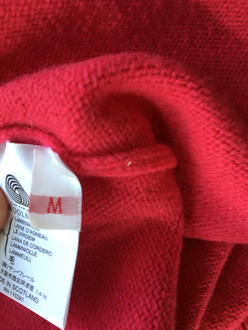May include: A white tag with black text attached to a red sweater. The tag says "WOOLN", "LAMBSW", "LAINE D'AGNEAU", "LA VIRGEM", "LANA DE CORDERO", "LAMMWOLLE", "LAMMEULL", "毛", "(株)サン・フレール", "大阪市西区阿波座 1-4-16", "DE IN SCOTLAND", "RN 110281". The tag also has a size "M" printed on it.