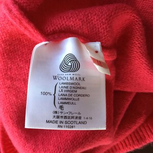 May include: A white tag with black text on a red sweater. The tag says "PURE NEW WOOL WOOLMARK LAMBSWOOL LAINE D'AGNEAU LA VIRGEM LANA DE CORDERO LAMMWOLLE LAMMEULL 毛 (株)サン・フレール 大阪市西区阿波座 1-4-16 MADE IN SCOTLAND RN 110281"