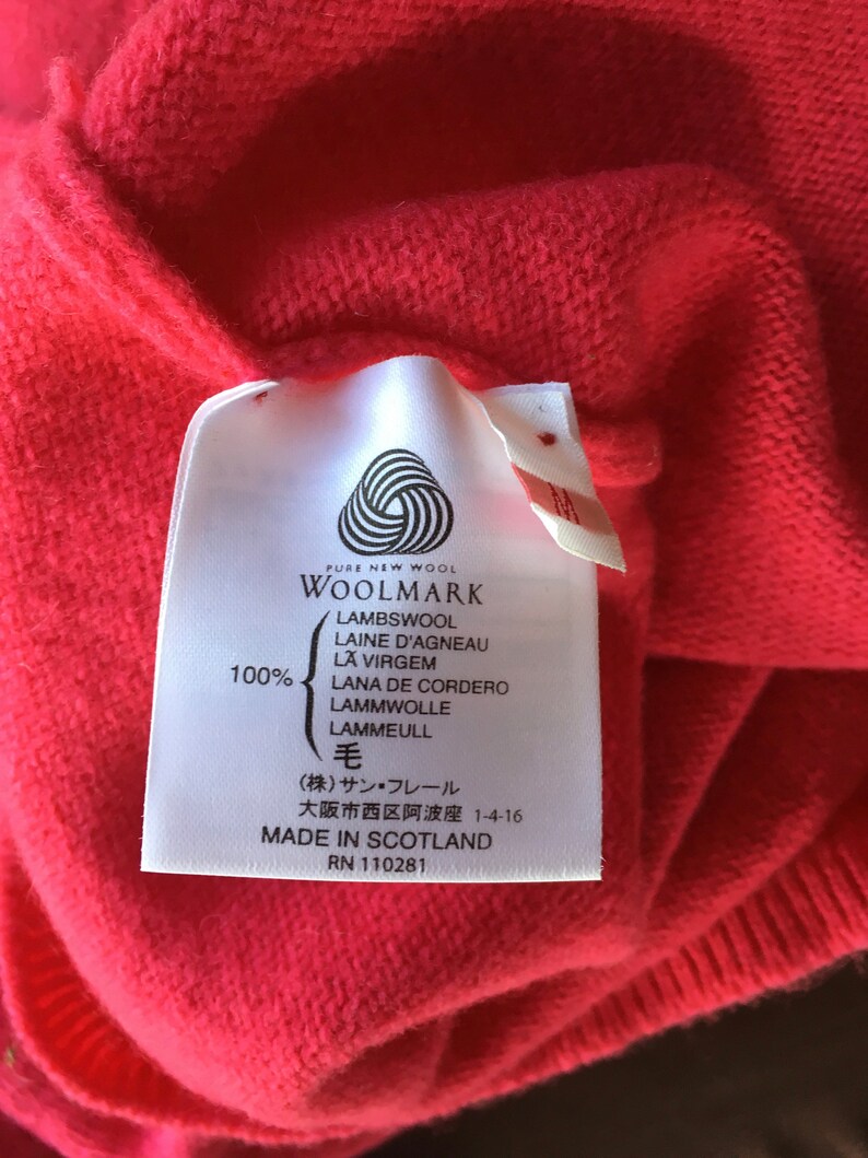 May include: A white tag with black text on a red sweater. The tag says "PURE NEW WOOL WOOLMARK LAMBSWOOL LAINE D'AGNEAU LA VIRGEM LANA DE CORDERO LAMMWOLLE LAMMEULL 毛 (株)サン・フレール 大阪市西区阿波座 1-4-16 MADE IN SCOTLAND RN 110281"