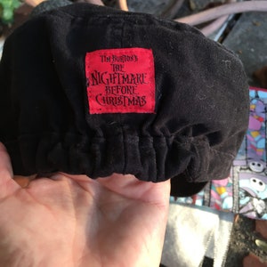 Op de afbeelding: Zwarte katoenen pet met een rood label waarop "Tim Burton's The Nightmare Before Christmas" staat.