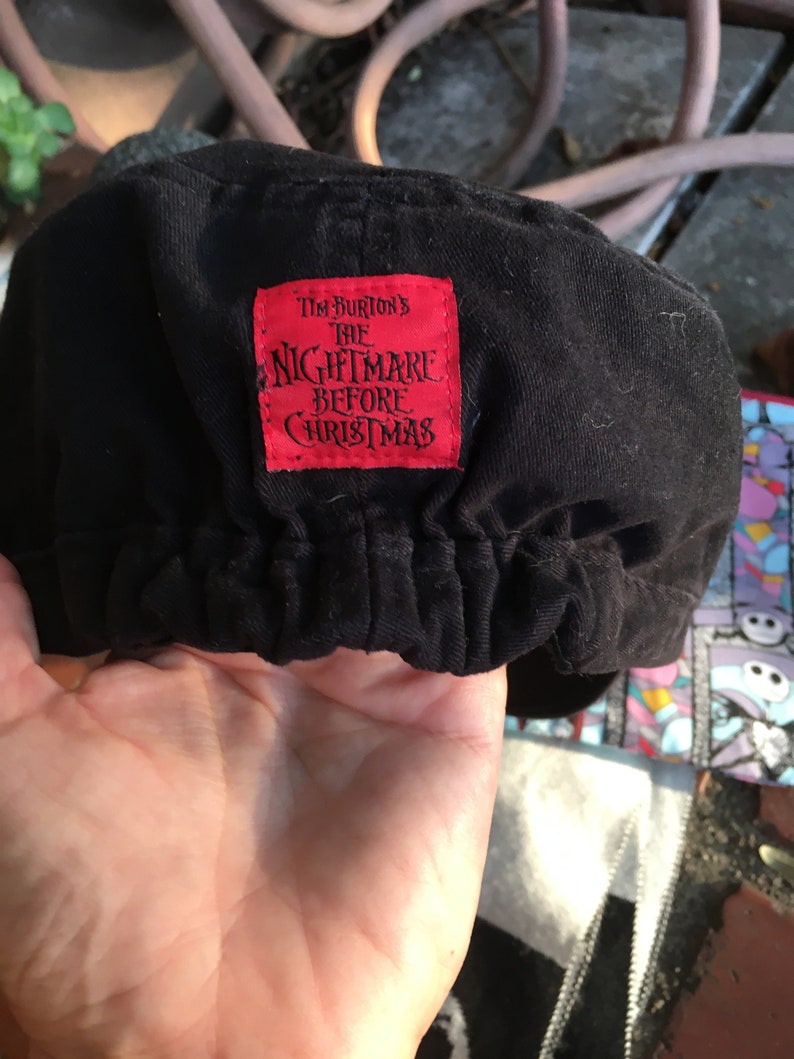 Op de afbeelding: Zwarte katoenen pet met een rood label waarop "Tim Burton's The Nightmare Before Christmas" staat.
