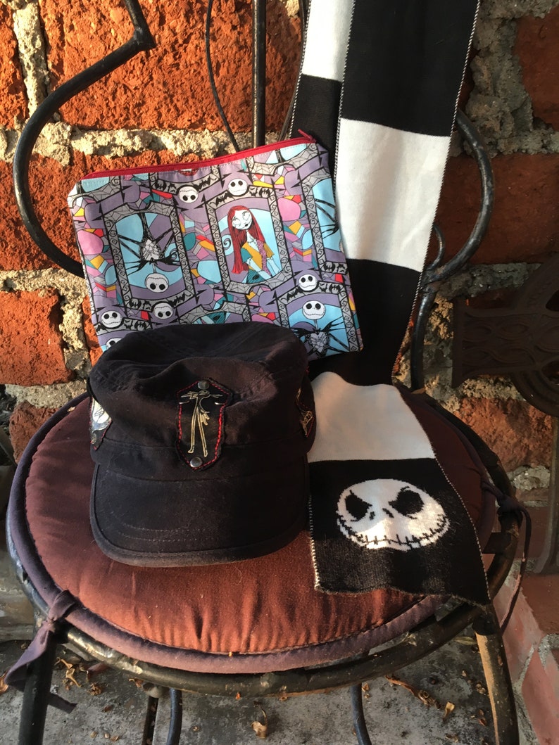 Op de afbeelding: Een zwart-wit gestreepte sjaal met een zwart-wit gebreid Jack Skellington gezicht aan de onderkant. Een zwarte pet met een rood en gouden geborduurd ontwerp. Een blauwe en paarse ritszak met een kleurrijk patroon van Jack Skellington en Sally.