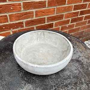 Puede incluir: Un cuenco de hormigón blanco con un interior liso y redondeado. El cuenco está sobre una superficie gris oscuro.