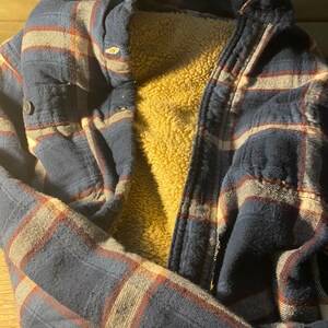 May include: A blue and red plaid flannel shirt with a brown fleece lining. The shirt is folded and laid flat on a brown surface.
