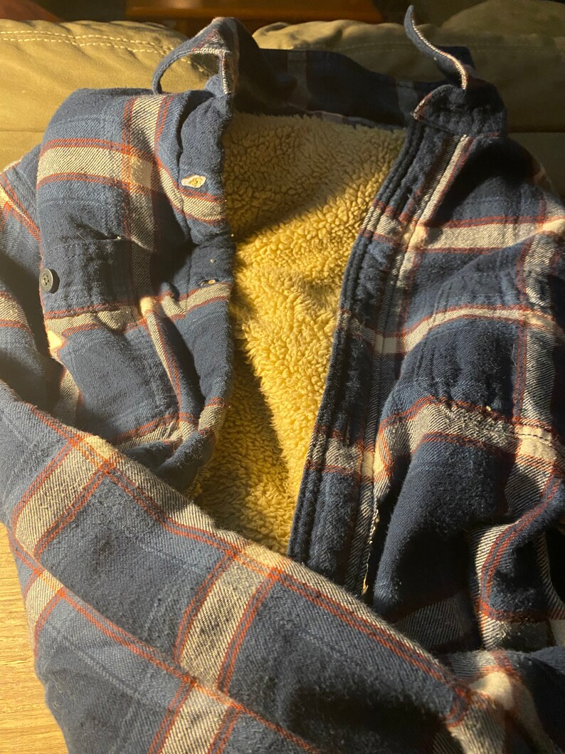 May include: A blue and red plaid flannel shirt with a brown fleece lining. The shirt is folded and laid flat on a brown surface.