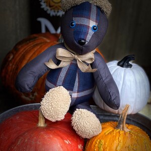 May include: A blue and brown plaid teddy bear with blue eyes sits on top of three pumpkins. The pumpkins are orange, red, and white.