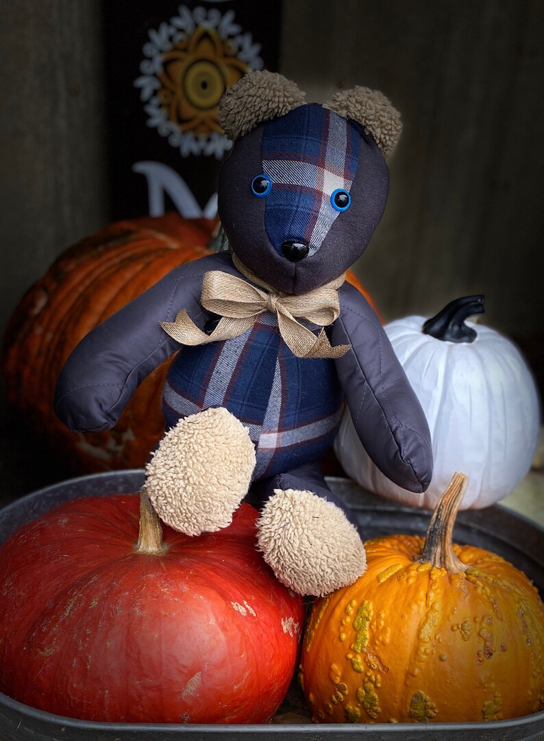 May include: A blue and brown plaid teddy bear with blue eyes sits on top of three pumpkins. The pumpkins are orange, red, and white.