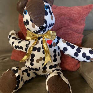May include: A white and black polka dot teddy bear with brown accents and blue eyes. The bear is wearing a gold ribbon and has a red heart pin on its chest. The bear is sitting on a red and brown couch.