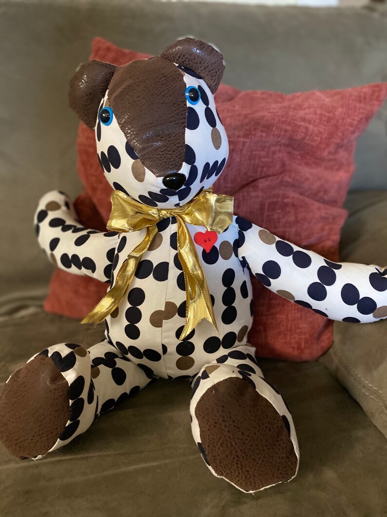 May include: A white and black polka dot teddy bear with brown accents and blue eyes. The bear is wearing a gold ribbon and has a red heart pin on its chest. The bear is sitting on a red and brown couch.