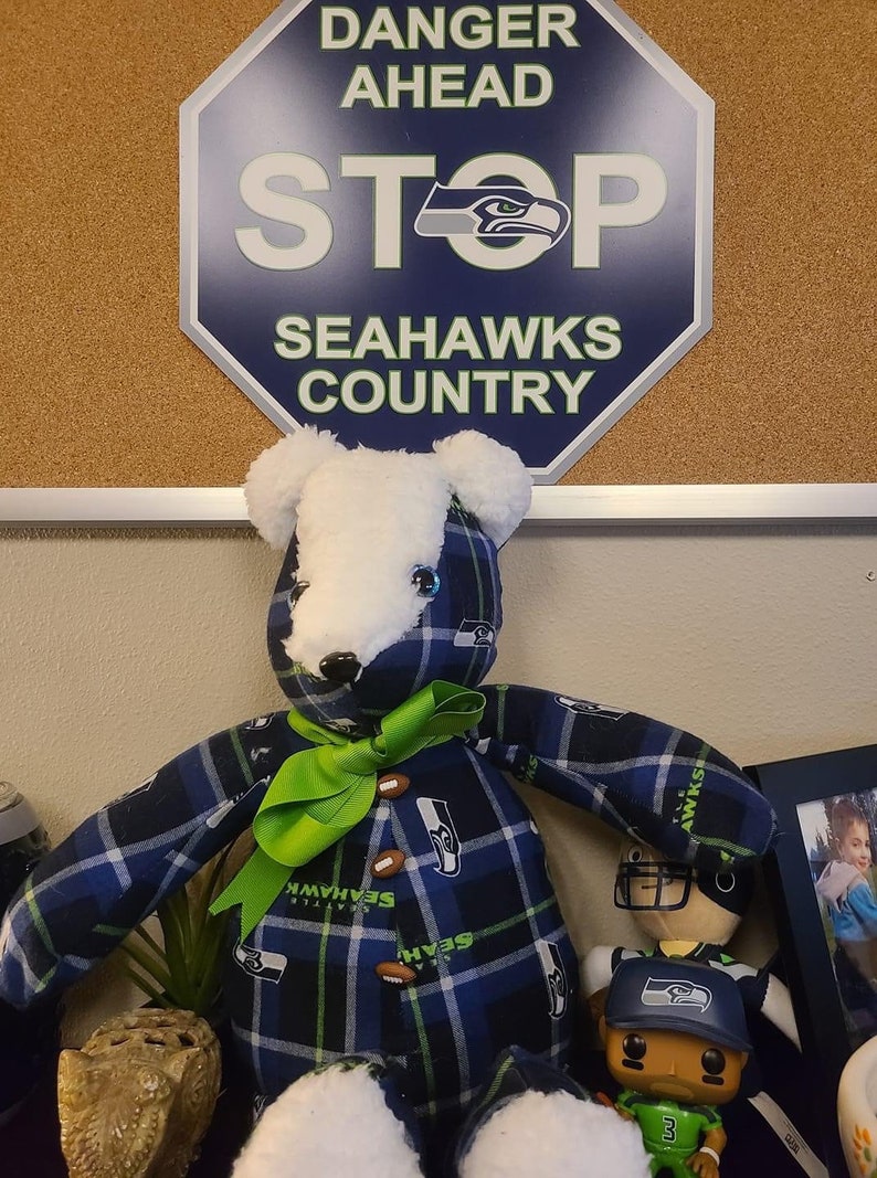 May include: A plush teddy bear wearing a blue and green plaid shirt with the Seattle Seahawks logo. The bear has a green bow and is sitting next to a small figurine of a football player wearing a Seahawks jersey and hat. The background is a corkboard with a sign that says "Danger Ahead Stop Seahawks Country".