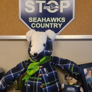 May include: A plush teddy bear wearing a blue and green plaid shirt with the Seattle Seahawks logo. The bear has a green bow and is sitting next to a small figurine of a football player wearing a Seahawks jersey and hat. The background is a corkboard with a sign that says "Danger Ahead Stop Seahawks Country".