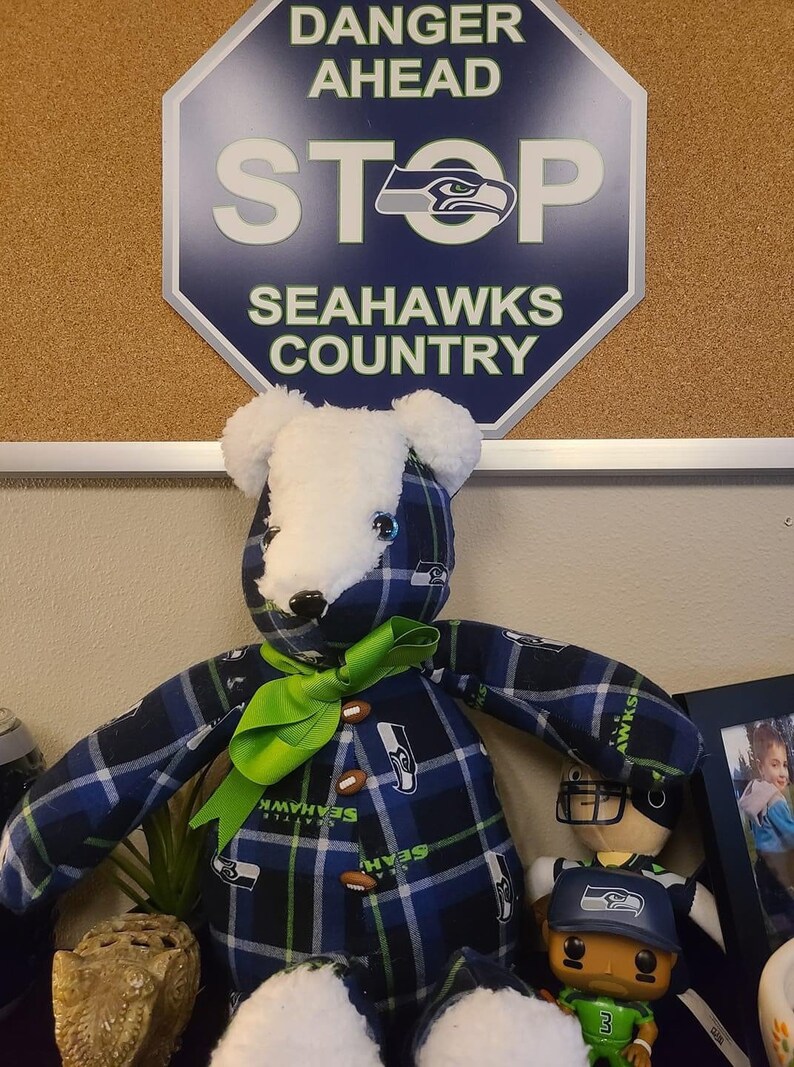 May include: A plush teddy bear wearing a blue and green plaid shirt with the Seattle Seahawks logo. The bear has a green bow and is sitting next to a small figurine of a football player wearing a Seahawks jersey and hat. The background is a corkboard with a sign that says "Danger Ahead Stop Seahawks Country".