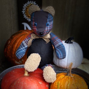 May include: A patchwork plaid teddy bear with blue and red accents sits on a table with pumpkins. The bear has black eyes and a black nose. The bear is wearing a brown ribbon around its neck.