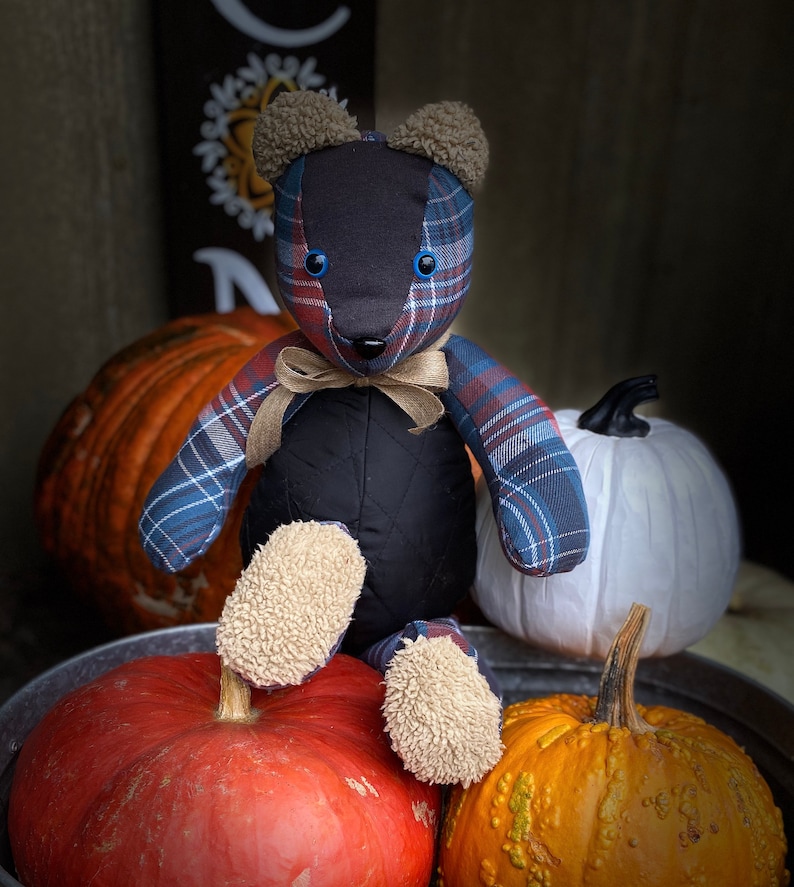 May include: A patchwork plaid teddy bear with blue and red accents sits on a table with pumpkins. The bear has black eyes and a black nose. The bear is wearing a brown ribbon around its neck.