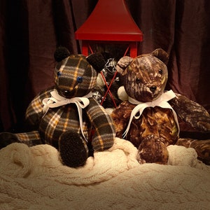 May include: Two plush teddy bears, one with a plaid pattern and the other with a camouflage pattern, are sitting on a white blanket in front of a red lantern. Both bears have white ribbon bows around their necks.