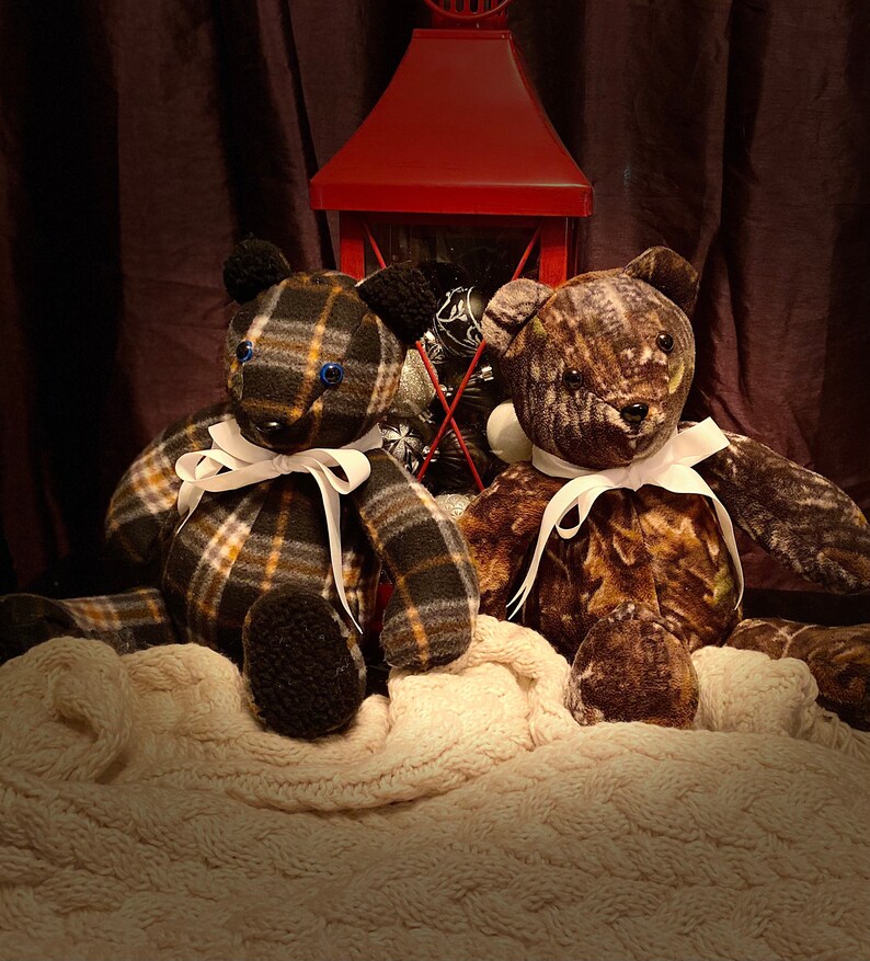 May include: Two plush teddy bears, one with a plaid pattern and the other with a camouflage pattern, are sitting on a white blanket in front of a red lantern. Both bears have white ribbon bows around their necks.
