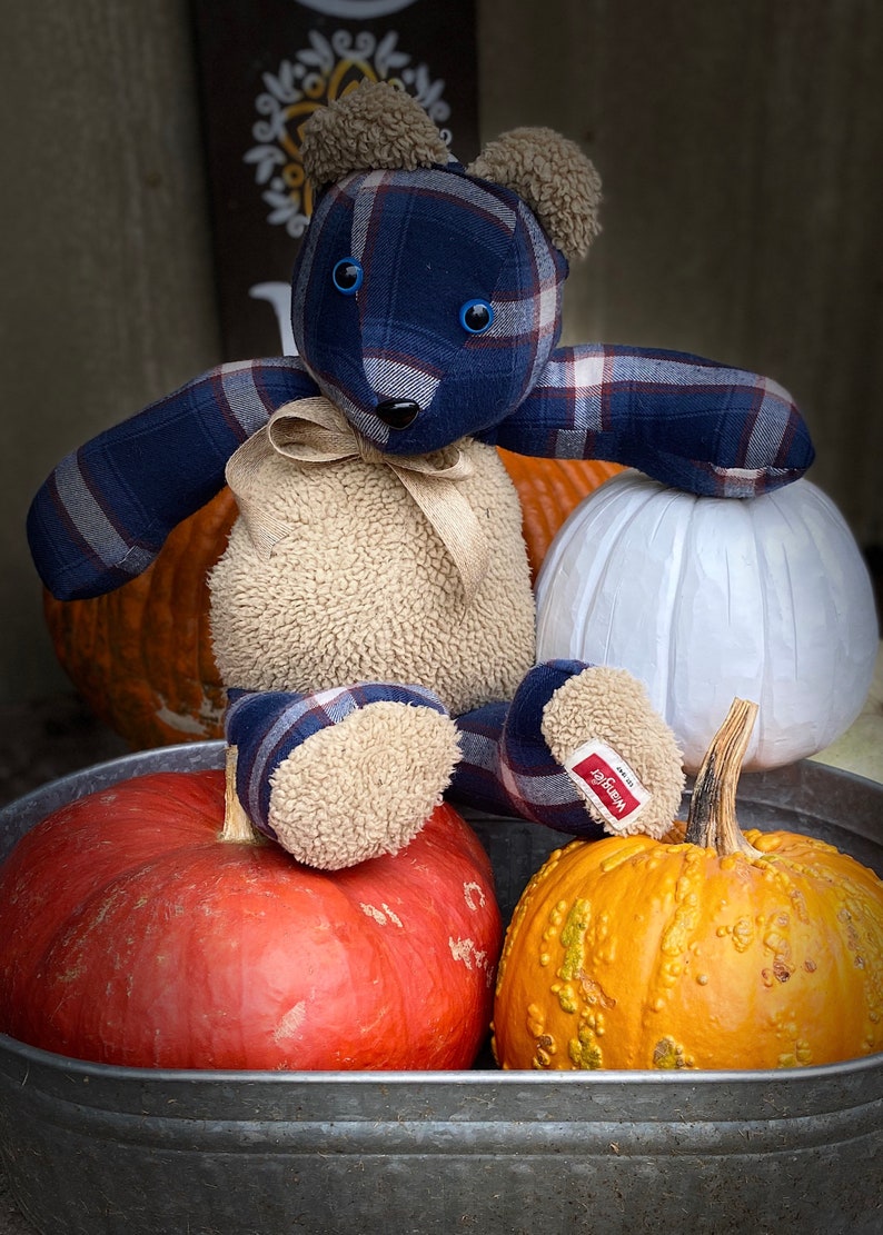 May include: A blue and white plaid teddy bear sitting on top of three pumpkins in a metal tub. The pumpkins are orange, red, and white. The teddy bear has blue eyes and a brown nose. The teddy bear is wearing a brown fur vest.