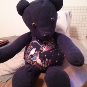 May include: A dark blue teddy bear with brown button eyes and a winter-themed fabric pattern on its chest. The bear is sitting on a wooden surface.